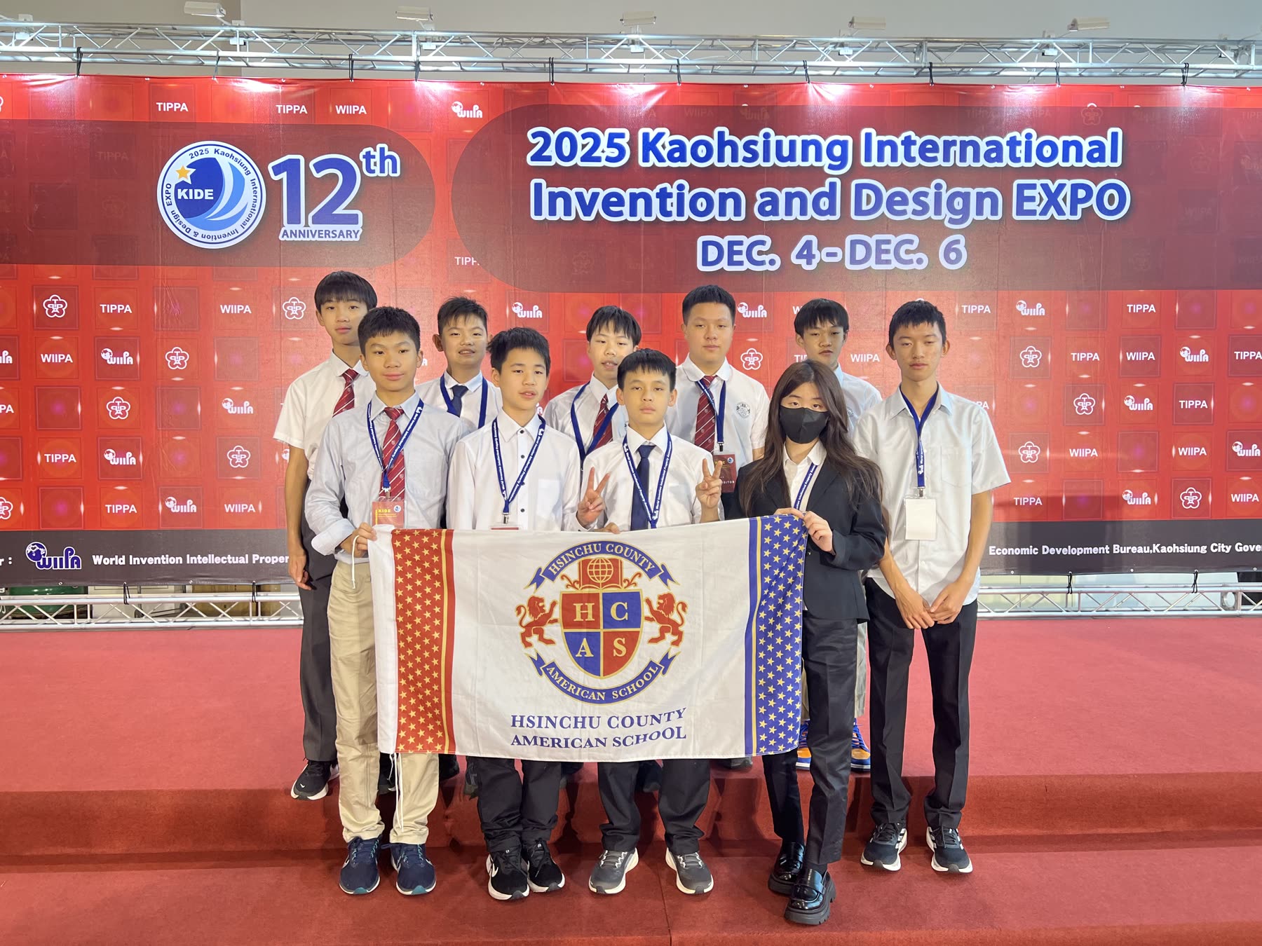 The HCAS team holding the school flag at the KIDE 2025 backdrop